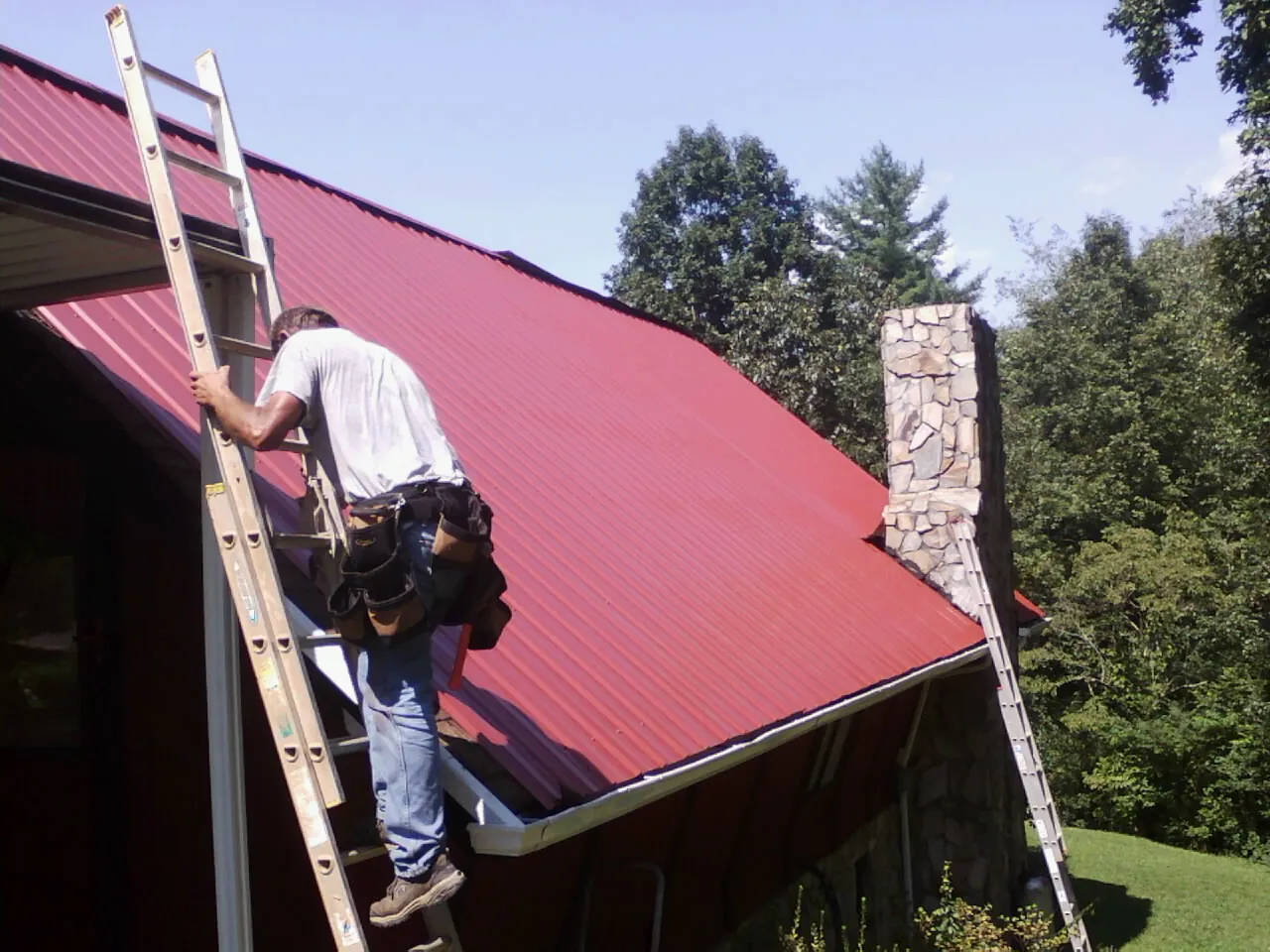 Roofer on a ladder beside a red metal roof and stone chimney — Soffit & Fascia Repair by Roofing Brothers in Shermans Dale